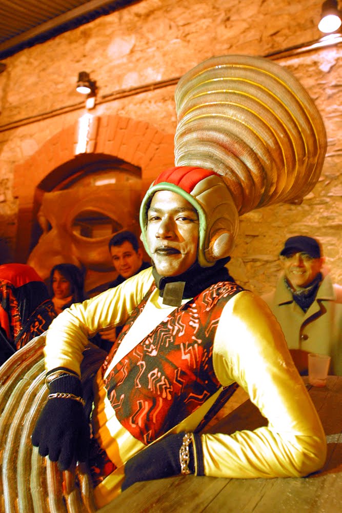 A close-up portrait of a performer in an elaborate, futuristic gold costume and oversized coiled headpiece, smiling warmly for the camera.